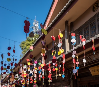 Colorful heart-shaped charms hang from wires in front of a traditional building. Each charm appears to have writing on it, suggesting they may be wishes or messages. The building features ornate wooden elements and a tiled roof. The sky is bright blue, enhancing the vibrant colors of the charms.