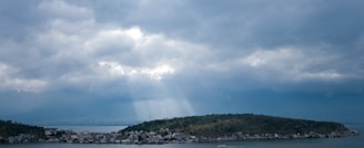 A small island is surrounded by water under a cloudy sky. Sunlight streams through breaks in the clouds, illuminating parts of the island. The island is covered with a mix of greenery and small buildings along its shoreline.