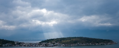 A small island is surrounded by water under a cloudy sky. Sunlight streams through breaks in the clouds, illuminating parts of the island. The island is covered with a mix of greenery and small buildings along its shoreline.