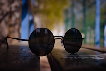 A pair of round, black sunglasses sit on a wooden surface. The background is softly blurred, featuring hints of green, blue, and brown, suggesting an outdoor setting. The overall atmosphere is tranquil, with subdued lighting and a shallow depth of field.