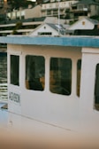 A close-up view of a boat with the registration number AGV993N, featuring a white exterior and blue roofing. The boat is docked near a waterfront with several modern houses in the background. The scene reflects off the calm water.