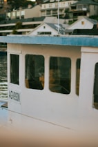 A close-up view of a boat with the registration number AGV993N, featuring a white exterior and blue roofing. The boat is docked near a waterfront with several modern houses in the background. The scene reflects off the calm water.