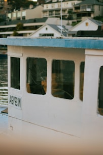 A close-up view of a boat with the registration number AGV993N, featuring a white exterior and blue roofing. The boat is docked near a waterfront with several modern houses in the background. The scene reflects off the calm water.