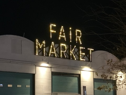 A night scene featuring a building with a large sign that reads 'FAIR MARKET' illuminated by numerous small lights. The building has a corrugated metal façade and two teal-colored doors. There are bare tree branches visible on the right side.