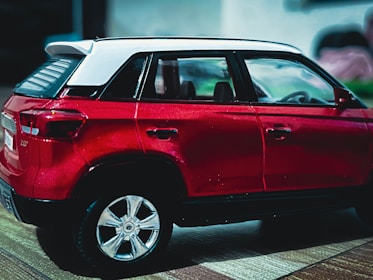 A detailed miniature model of a red SUV with a white roof is shown. The model car has intricate details, including visible side mirrors, door handles, and alloy wheels. The background is blurred, making the focus entirely on the car model. The car is positioned on a flat textured surface.