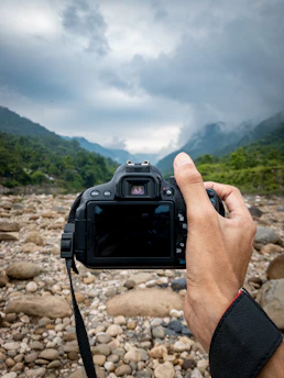 Traveler holding a DSLR camera against a scenic mountain backdrop.