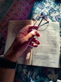 A hand holding glasses above an open book with text written in an Indian language, possibly Hindi. The book is placed on a colorful patterned fabric that covers a surface.