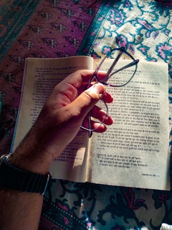 A hand holding glasses above an open book with text written in an Indian language, possibly Hindi. The book is placed on a colorful patterned fabric that covers a surface.