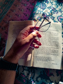 A hand holding glasses above an open book with text written in an Indian language, possibly Hindi. The book is placed on a colorful patterned fabric that covers a surface.