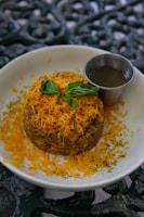 A round dish topped with finely grated orange cheese and garnished with fresh basil leaves rests on a white plate. Beside it, a small metal cup contains a dark sauce. The plate sits on an ornate, dark-colored surface.
