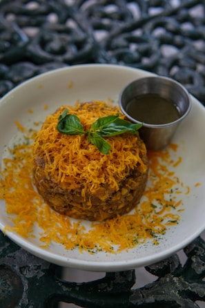 A round dish topped with finely grated orange cheese and garnished with fresh basil leaves rests on a white plate. Beside it, a small metal cup contains a dark sauce. The plate sits on an ornate, dark-colored surface.