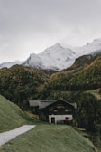 A rustic wooden cabin is nestled in a lush green valley surrounded by densely forested hills. In the background, snow-covered mountain peaks rise majestically against a cloudy sky, providing a serene and picturesque alpine scene.