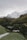 A rustic wooden cabin is nestled in a lush green valley surrounded by densely forested hills. In the background, snow-covered mountain peaks rise majestically against a cloudy sky, providing a serene and picturesque alpine scene.