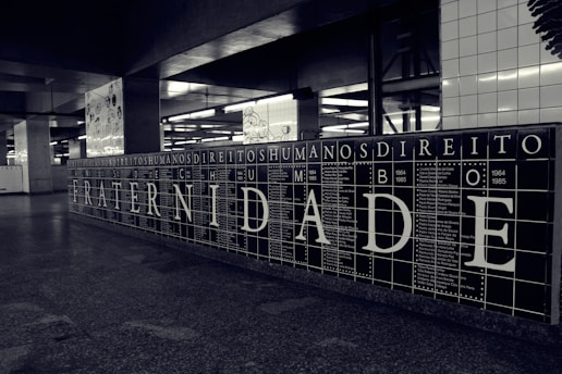 A black and white tiled wall displays large letters spelling out 'FRATERNIDADE,' which means brotherhood in Portuguese. Surrounding the main word are smaller words and years, possibly representing historical events or names. The setting appears to be an underground station or public space with additional artwork visible in the background.