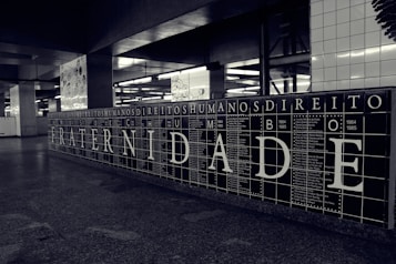 A black and white tiled wall displays large letters spelling out 'FRATERNIDADE,' which means brotherhood in Portuguese. Surrounding the main word are smaller words and years, possibly representing historical events or names. The setting appears to be an underground station or public space with additional artwork visible in the background.