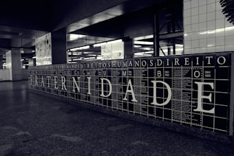 A black and white tiled wall displays large letters spelling out 'FRATERNIDADE,' which means brotherhood in Portuguese. Surrounding the main word are smaller words and years, possibly representing historical events or names. The setting appears to be an underground station or public space with additional artwork visible in the background.