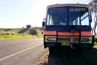 A colorful, retro-style bus is parked on the side of a rural road. The bus has a painted message on its front window that reads 'BE HAPPY.' The setting features a sunny, open landscape with green fields and a few scattered trees. The road is seemingly quiet, giving a sense of calm and tranquility.