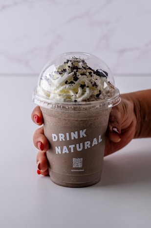 A hand with red-painted nails holds a chocolate smoothie topped with whipped cream and chocolate crumbles, in a clear plastic cup with the words 'DRINK NATURAL' printed on it. The background is a simple, light-colored surface.