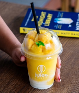 A hand holding a vibrant yellow smoothie in a clear plastic cup with a lid, topped with chunks of fruit and a fresh mint leaf, accompanied by a black straw. In the background, a book with a blue and yellow cover is partially visible on a wooden table.