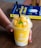 A hand holding a vibrant yellow smoothie in a clear plastic cup with a lid, topped with chunks of fruit and a fresh mint leaf, accompanied by a black straw. In the background, a book with a blue and yellow cover is partially visible on a wooden table.