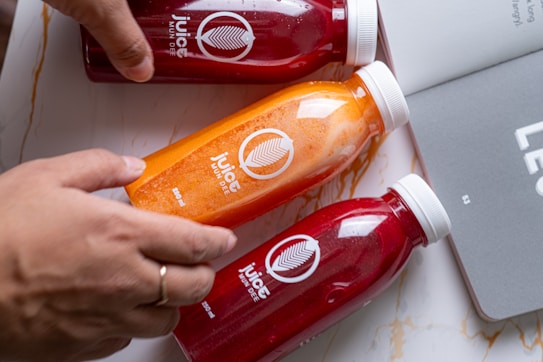 Three bottles of juice with bright colored liquids inside are placed on a marble surface next to a gray notebook. Two bottles contain a red liquid, and one contains an orange liquid. A hand with a ring is holding the orange juice bottle. The bottles have white caps and labels with the logo and text.
