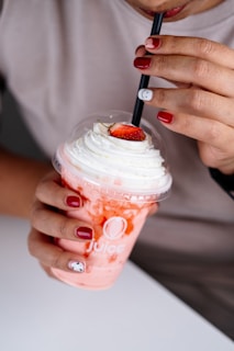 A person is sipping a strawberry smoothie topped with whipped cream and a sliced strawberry from a plastic cup with a logo. The person has neatly painted red and white nails, with an artistic design on one of the nails.