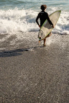 A surfer wearing Salvage Surf apparel walking towards the ocean with a skeleton-themed surfboard under arm.