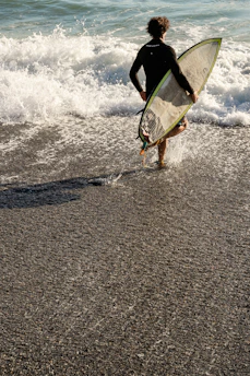 A surfer wearing Salvage Surf apparel walking towards the ocean with a skeleton-themed surfboard under arm.