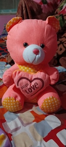 A plush pink teddy bear with orange and white polka dot accents on its ears and feet is seated on a colorful patterned surface. The bear holds a heart with the words 'With Love' printed on it. The background includes a brown blanket with floral design.