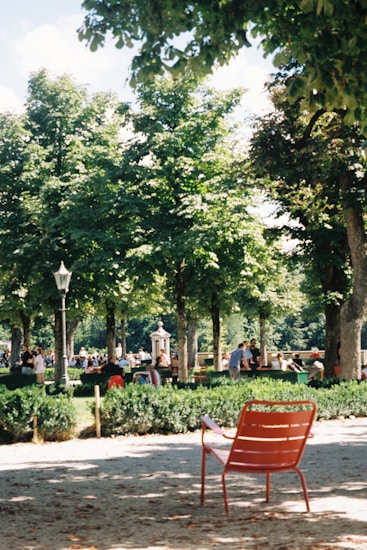 A park scene featuring a red metal chair in the foreground on a sandy path. The background is filled with lush green trees and numerous people engaging in leisure activities, creating a sense of community and relaxation. A lamppost and a decorative structure are also visible, adding to the park's ambiance.