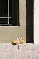 A single tan-colored shoe with white shoelaces is placed on a concrete ledge. The shoe appears worn and is situated in front of a window with metal bars. The wall behind the shoe has a rough texture, and the scene is brightly lit by sunlight, creating a contrast between the shadowed and lit areas.