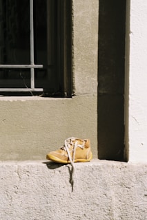 A single tan-colored shoe with white shoelaces is placed on a concrete ledge. The shoe appears worn and is situated in front of a window with metal bars. The wall behind the shoe has a rough texture, and the scene is brightly lit by sunlight, creating a contrast between the shadowed and lit areas.