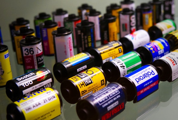 A collection of colorful 35mm film canisters is arranged in various rows. The canisters feature different brands and ISO numbers, showcasing the variety of film types available. Text and logos in various fonts are visible across the canisters, indicating brand names and film specifications.