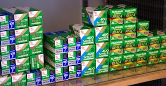 A collection of Fujifilm photographic film boxes, neatly stacked with labels such as Sensia II, Provia, and Fujicolor Superia Reala. The boxes are primarily green and white with some blue and red accents, arranged on a reflective surface.