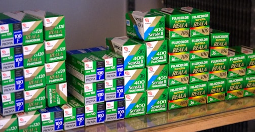 A collection of Fujifilm photographic film boxes, neatly stacked with labels such as Sensia II, Provia, and Fujicolor Superia Reala. The boxes are primarily green and white with some blue and red accents, arranged on a reflective surface.
