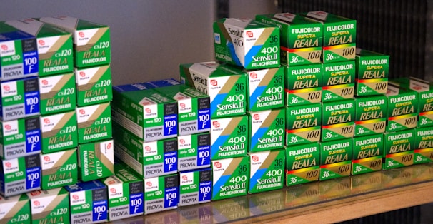 A collection of Fujifilm photographic film boxes, neatly stacked with labels such as Sensia II, Provia, and Fujicolor Superia Reala. The boxes are primarily green and white with some blue and red accents, arranged on a reflective surface.
