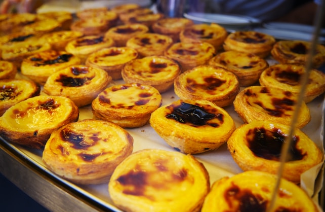 A display of multiple freshly baked tarts, each with a golden-brown crispy crust and a smooth custard filling, some with a slightly burnt top. The tarts are arranged closely together on a baking tray.