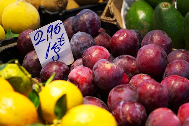 A market display of fresh fruits, including several plums in the foreground, with a price tag showing the cost per kilogram in Euros. There are also some lemons and avocados visible around the plums.