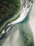 A winding road runs parallel to a coastline, bordered on one side by dense green forest and on the other by white sandy beach and clear turquoise water. The area appears serene and untouched, offering a blend of rugged natural beauty and tranquility.