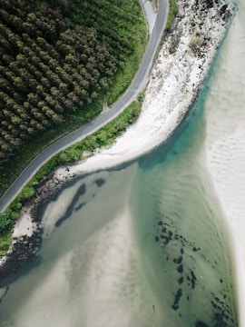A winding road runs parallel to a coastline, bordered on one side by dense green forest and on the other by white sandy beach and clear turquoise water. The area appears serene and untouched, offering a blend of rugged natural beauty and tranquility.