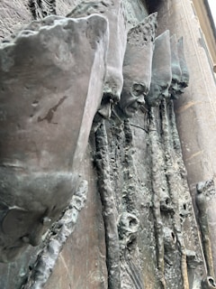 A detailed view of a textured metal sculpture featuring elongated, abstract human faces with tall, pointed hats, arranged in a row. The sculpture has a rough, aged surface with intricate details and a patina finish that adds depth and shadows.