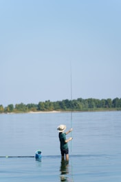 A person is standing waist-deep in a calm body of water while holding a fishing rod. They are wearing a wide-brimmed hat and are casting a line into the water. In the distance, a line of trees separates the water from the clear blue sky. A blue fishing basket floats nearby.