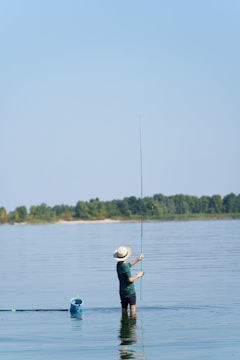 A person is standing waist-deep in a calm body of water while holding a fishing rod. They are wearing a wide-brimmed hat and are casting a line into the water. In the distance, a line of trees separates the water from the clear blue sky. A blue fishing basket floats nearby.