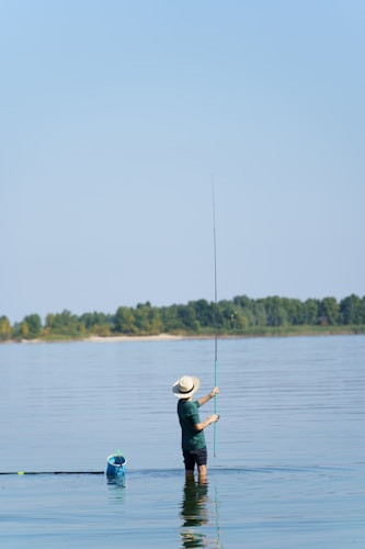 A person is standing waist-deep in a calm body of water while holding a fishing rod. They are wearing a wide-brimmed hat and are casting a line into the water. In the distance, a line of trees separates the water from the clear blue sky. A blue fishing basket floats nearby.