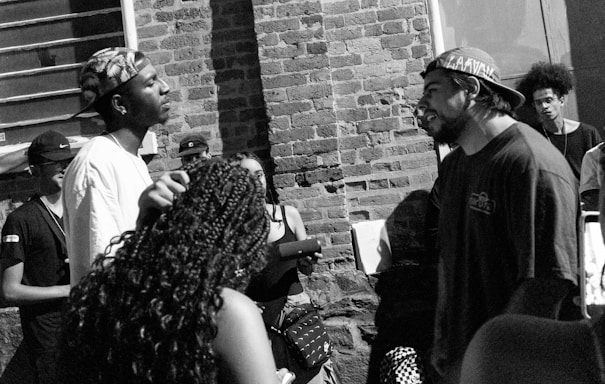 A group of people gather outdoors near a brick wall, engaged in what appears to be a conversation or rap battle. Two individuals standing face-to-face seem to be the focal point, surrounded by onlookers. The image is in black and white, adding a gritty, urban feel.