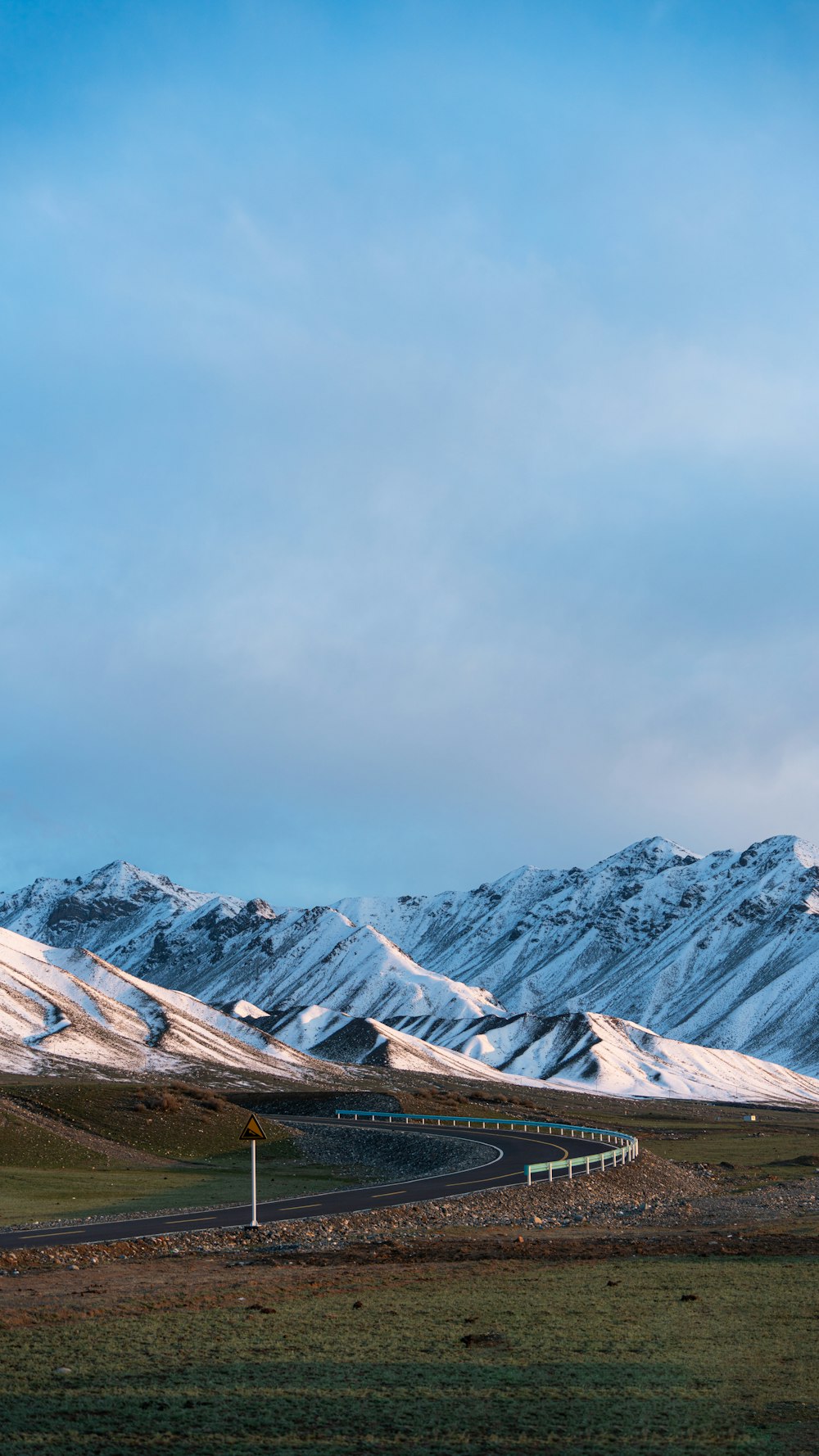 a road in the middle of a field with snow covered mountains in the background