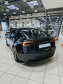 A sleek, black electric car is positioned on a tiled showroom floor. The modern interior of the showroom features large windows, numerous overhead lights, and industrial-style metal ceiling structures. Other vehicles and showroom elements can be seen in the background.