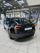 A sleek, black electric car is positioned on a tiled showroom floor. The modern interior of the showroom features large windows, numerous overhead lights, and industrial-style metal ceiling structures. Other vehicles and showroom elements can be seen in the background.