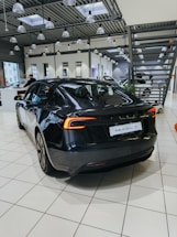 A sleek, black electric car is positioned on a tiled showroom floor. The modern interior of the showroom features large windows, numerous overhead lights, and industrial-style metal ceiling structures. Other vehicles and showroom elements can be seen in the background.