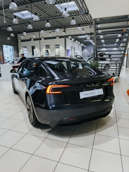 A sleek, black electric car is positioned on a tiled showroom floor. The modern interior of the showroom features large windows, numerous overhead lights, and industrial-style metal ceiling structures. Other vehicles and showroom elements can be seen in the background.